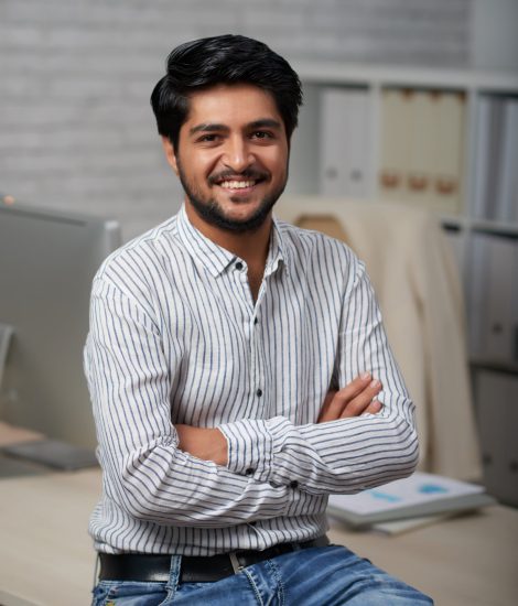 Confident young Indian businessman sitting on edge of his table and looking at camera